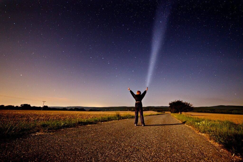 person raising his two hands while holding flashlight standing on road between grass during sunset